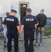 policjanci  przed budynkiem komendy stoja z dwoma zatrzymanymi