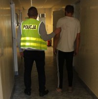 policjant Wydziału Kryminalnego w kamizelce z napisem POLICJA prowadzi zatrzymanego mężczyznę.