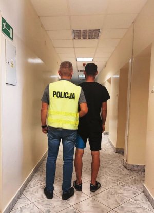 policjant w kamizelce z napisem POLICJA prowadzi zatrzymanego.