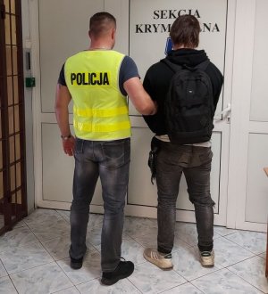 policjant w kamizelce z napisem POLICJA prowadzi zatrzymanego.