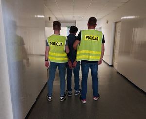 policjanci w kamizelkach z napisem POLICJA prowadzą zatrzymanego.