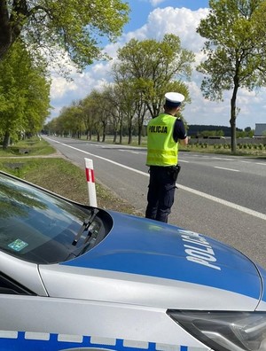 policjant kontroluje prędkość pojazdów na drodze, w tle policyjny radiowóz.