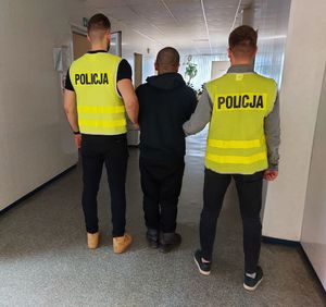 policjanci w kamizelkach z napisem POLICJA prowadzi zatrzymanego.