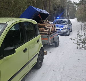 radiowóz policji na sygnałach a przed nim samochód z przyczepka wypełnioną drewnem.