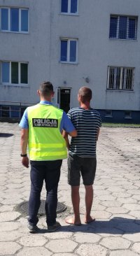 policjant w kamizelce odblaskowej z napisem policji stoi na terenie zaplecza komendy z zatrzymanym.stoja tyłem